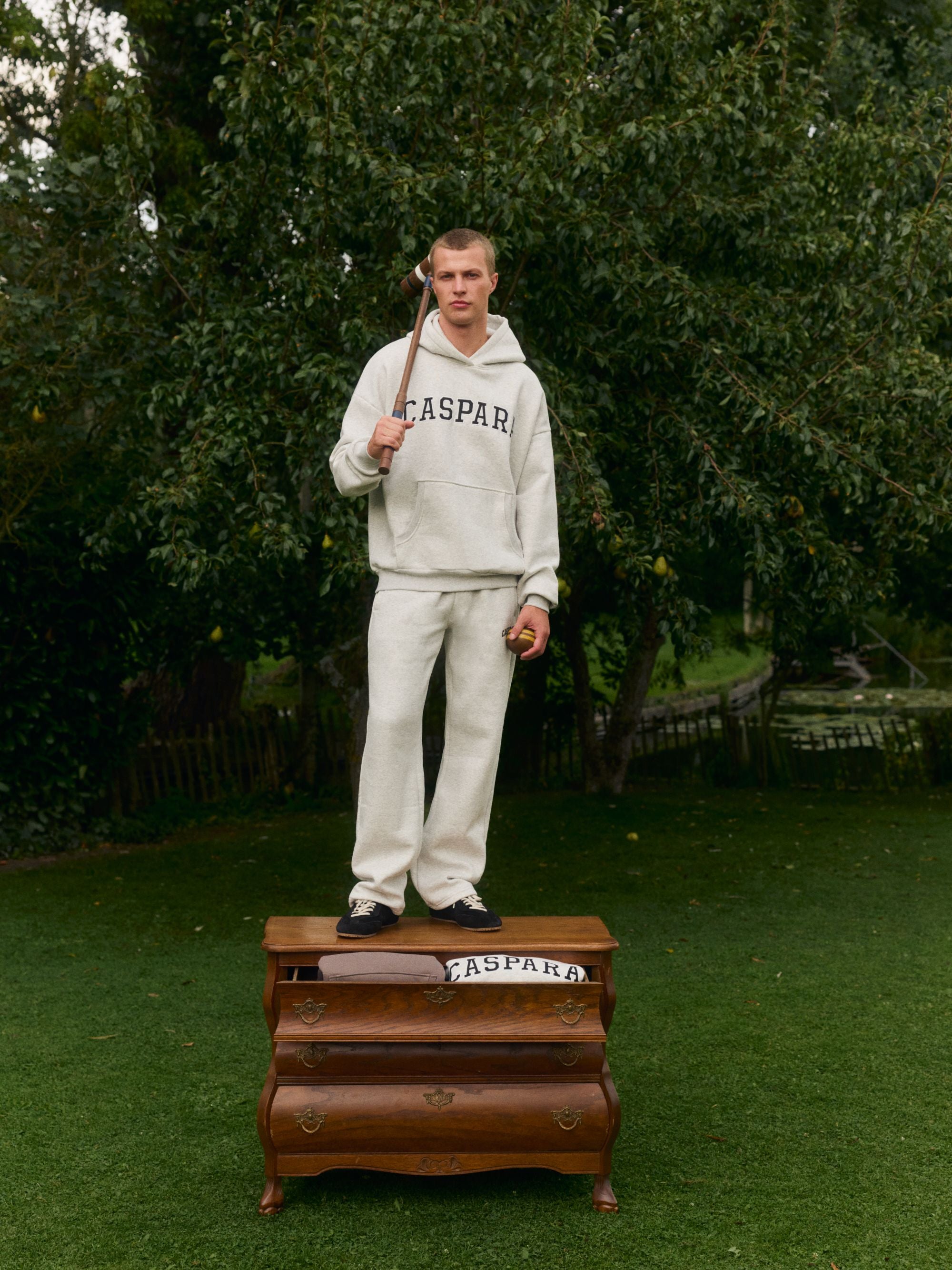 Man wearing a grey melange CASPARA logo hoodie and matching sweatpants standing outdoors on a wooden chest.